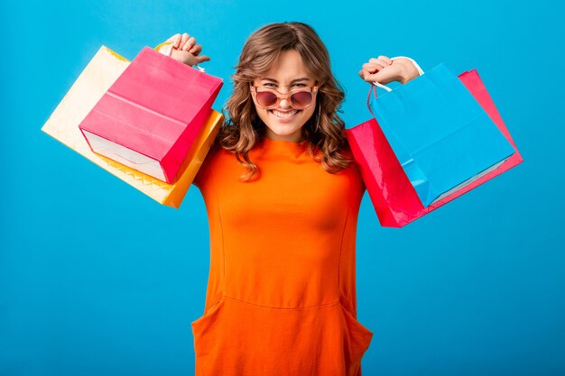 Portrait of excited attractive smiling stylish woman shopaholic in orange trendy dress holding shopping bags on blue studio background isolated