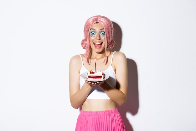 Портрет взволнованной именинницы в розовом парике, которая выглядит удивленной, загадывая желание на торте с зажженной свечой, празднуя.