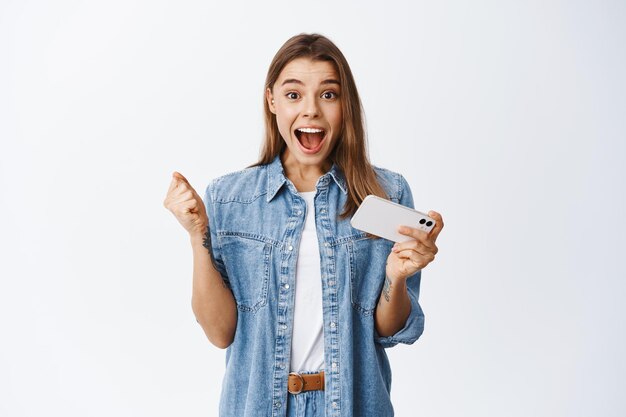 Portrait of excited blond girl winning money or prize online, holding mobile phone and screaming for joy, celebrating victory, winning in video game on smartphone, white