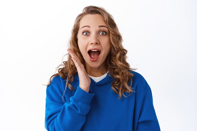 Portrait of excited blond woman gasping, drop jaw and looking impressed at front, checking out awesome special promo offer, standing over white wall