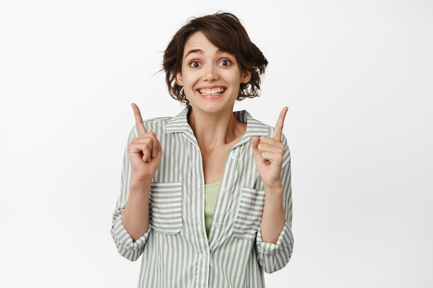 笑顔で興奮しているブルネットの少女の肖像画、左右に指を指して、白い背景の上に立って、幸せな喜びの顔で広告を表示します。