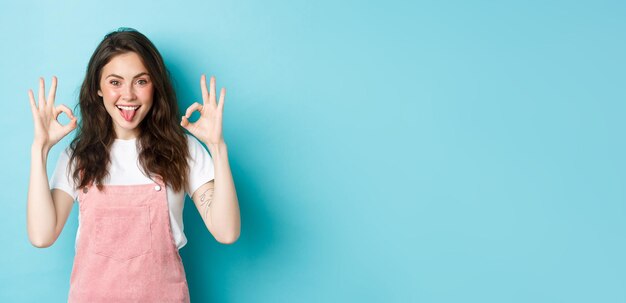 Ritratto di ragazza bruna eccitata con acconciatura riccia che mostra segni ok elogiare scelta perfetta consigliare cosa buona dare segno ok in piedi su sfondo blu