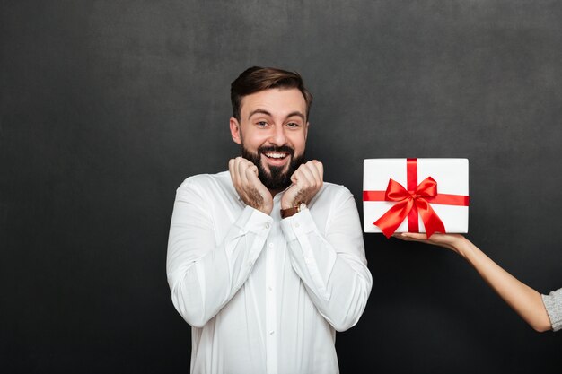 Portrait of excited brunette man rejoicing to get white gift box with red bow from female hand over dark gray wall