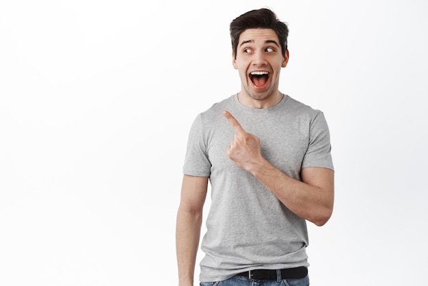 Portrait of excited caucasian male model pointing aside and scream from joy and amazement standing over white background