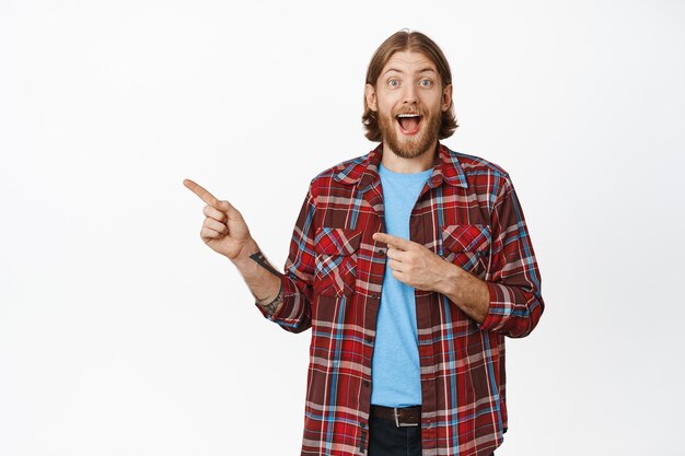 Portrait of excited caucasian man with beard and happy smiling face, pointing fingers left, showing amazing discount sale, big promo, standing over white background