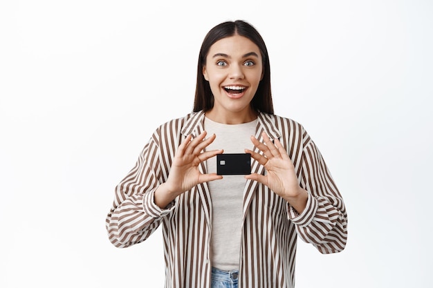 Portrait of excited caucasian woman in stylish outfit, showing plastic credit card and smiling amazed, standing over white wall