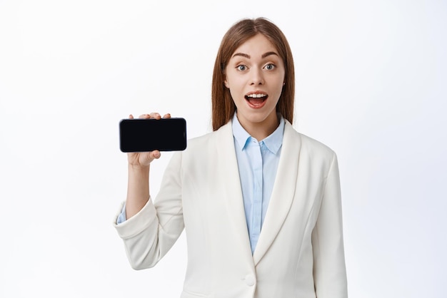 Портрет взволнованной корпоративной девушки-офисного работника в костюме показывает пустой экран смартфона и изумленную улыбку демонстрирует приложение или веб-сайт на белом фоне
