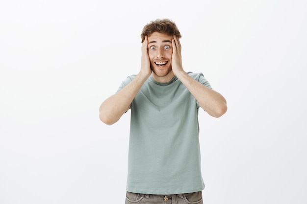 Portrait of excited crazy european guy in t-shirt, holding hands on face and smiling with weird funny expression