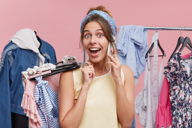 Free photo portrait of excited female talking over smart phone while holding hangers with clothes in hand, looking with happiness and surprise