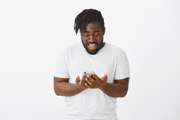 Portrait of excited guy with braids posing against the white wall with his phone