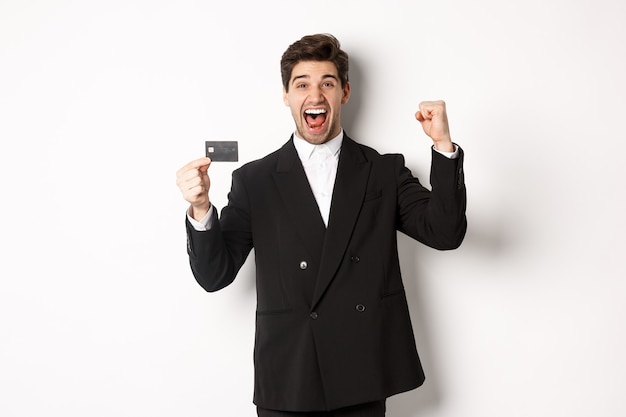 白い背景に立って、喜んでクレジットカードを表示し、スーツを着て興奮しているハンサムなビジネスマンの肖像画。