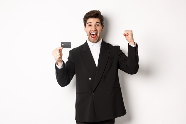 Портрет возбужденного красивого бизнесмена в костюме, радуясь и показывая кредитную карту, стоя на белом фоне.
