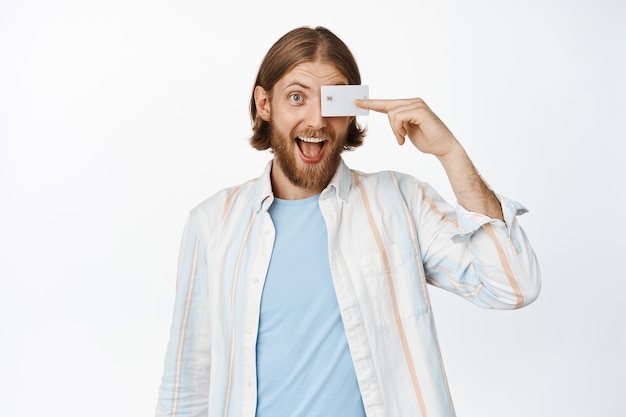 Portrait of excited handsome guy looking happy, holding credit discount card on eye, standing on white. Shopping and money concept