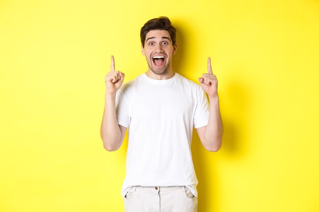 Portrait of excited handsome man in white t-shirt, pointing fingers up, showing offer, standing against yellow background.