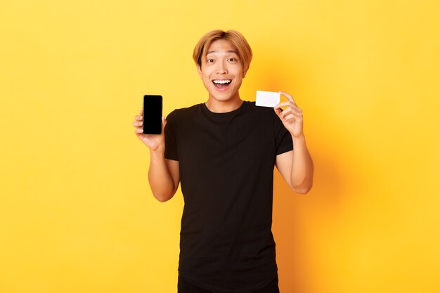 Portrait of excited happy asian man showing mobile phone screen and credit card with joyful smile, standing yellow wall