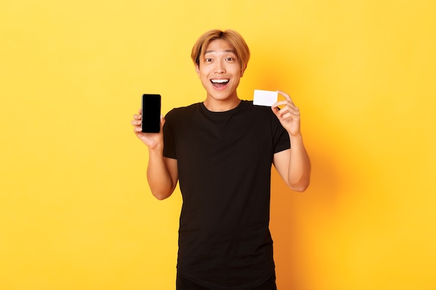Портрет возбужденного счастливого азиатского мужчины, показывающего экран мобильного телефона и кредитную карту с радостной улыбкой, стоящего на желтой стене