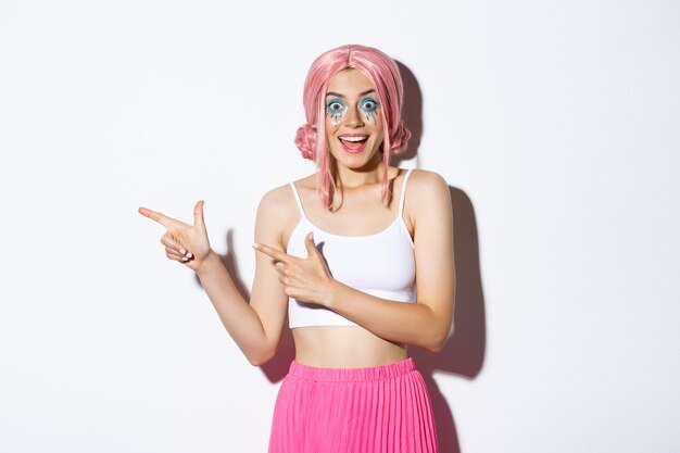 Portrait of excited happy girl in pink wig and bright makeup celebrating something, pointing fingers left at your logo about holidays and parties, standing over white background