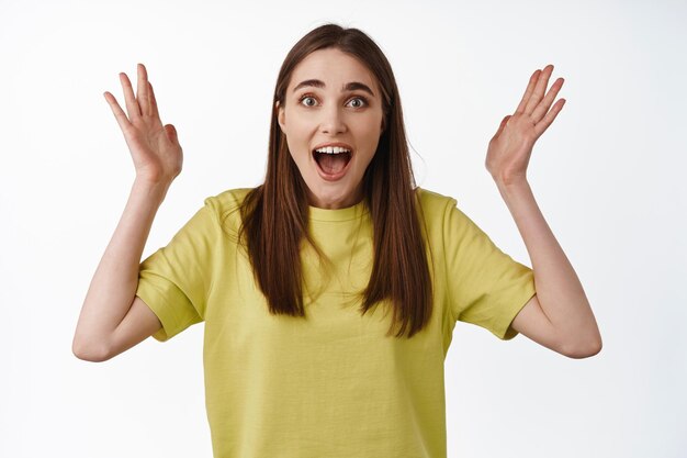 Portrait of excited happy woman, gasping astonished, raising hands up celebrating, surprised to hear something awesome, standing over white background
