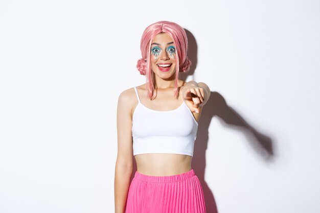 Portrait of excited happy young girl in pink wig and halloween costume, pointing finger at camera