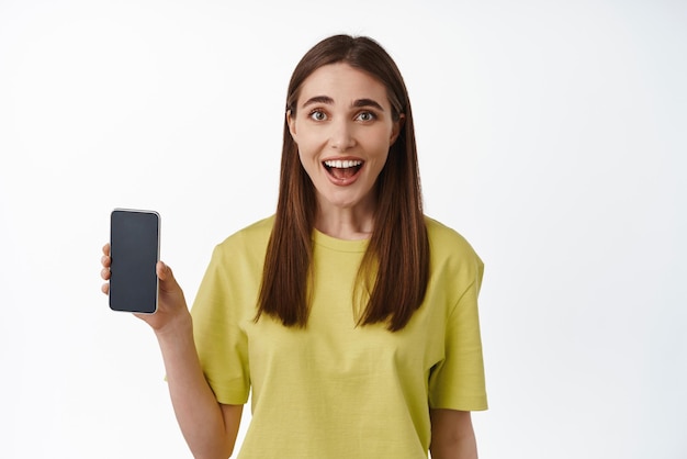 Portrait of excited lucky girl shows smartphone screen app interface smiling amazed recommending shopping application standing against white background