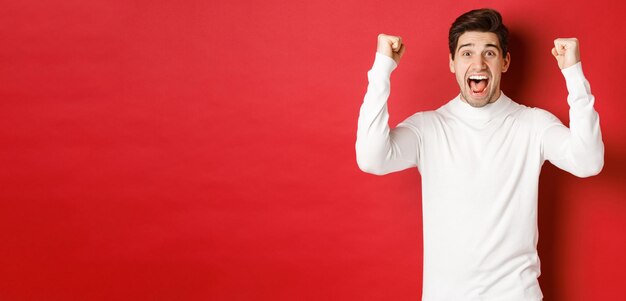 Portrait of excited lucky man in white sweater raising hands up and triumphing celebrating new year standing happy against red background