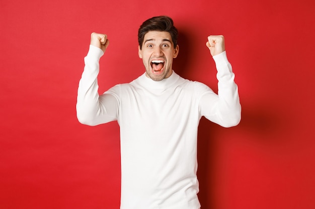 Portrait of excited lucky man in white sweater raising hands up and triumphing celebrating new year ...