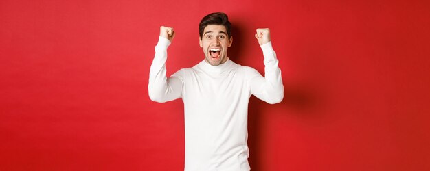 Portrait of excited lucky man in white sweater raising hands up and triumphing celebrating new year ...