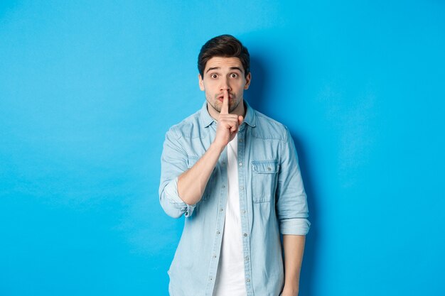 Portrait of excited man asking to keep quiet, showing hush taboo sign and looking nervously at camera, standing against blue background