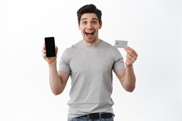 Portrait of excited man showing empty phone screen and credit card demonstrate online store and making order standing over white background