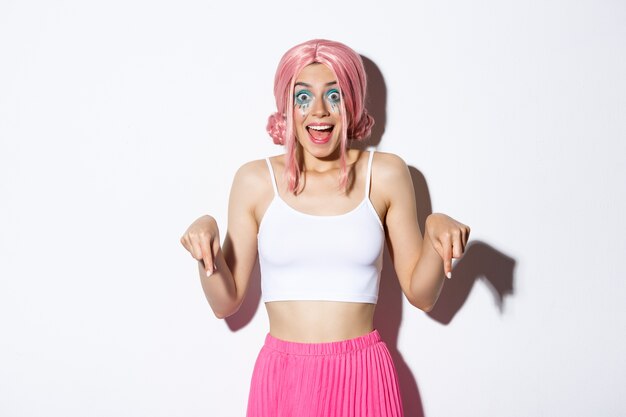 Portrait of excited party girl in pink wig and halloween outfit, looking amused and happy while pointing fingers down, standing.