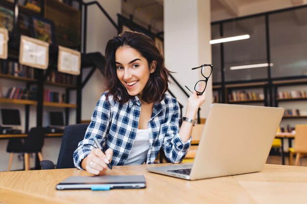 초상화 흥분 미소 갈색 머리 젊은 여자 도서관에서 노트북으로 작업. 영리한 학생, 대학 생활, 인터넷에서 일하고, 웃고, 쾌활한 분위기.