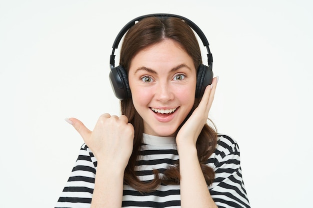 Free photo portrait of excited woman in wireless headphones showing awesome news pointing left standing over