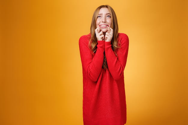 Portrait of excited and worried young redhead female in 20s stooping standing in begging pose crossing finger for good luck looking at sky as praying god and asking help with wish come true