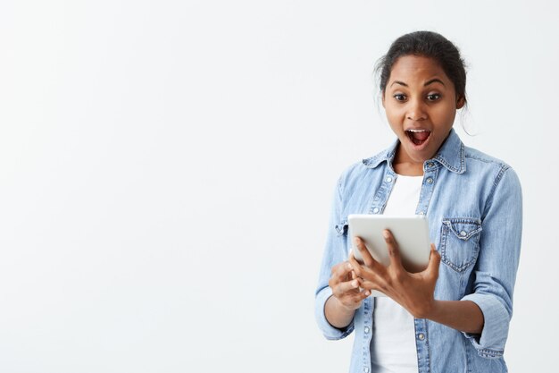 Portrait of excited young African American female screaming in shock and amazement holding new tablet in her hands. Surprised bug-eyed dark-skinned girl looking impressed, can't believe her own eyes a