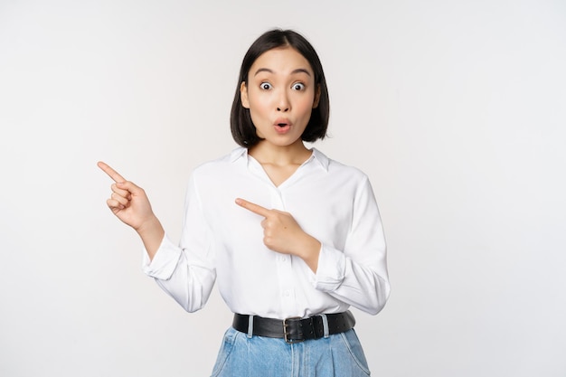 Portrait of excited young asian woman office lady pointing fingers left at discount showing sale banner standing over white background