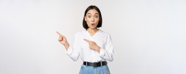 Portrait of excited young asian woman office lady pointing fingers left at discount showing sale banner standing over white background