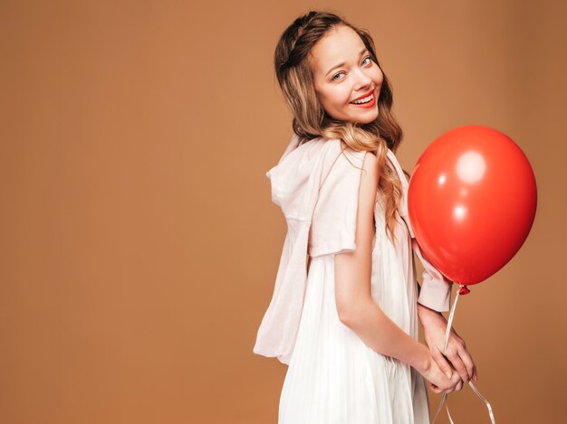 Портрет возбужденных молодая девушка позирует в модном летнем белом платье. Улыбающиеся женщина с красный шар позирует. Модель готова к вечеринке