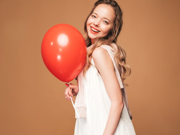 Портрет возбужденных молодая девушка позирует в модном летнем белом платье. Улыбающиеся женщина с красный шар позирует. Модель готова к вечеринке