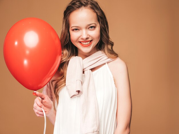 Portrait of excited young girl posing in trendy summer white dress. Smiling woman with red balloon posing. Model ready for party