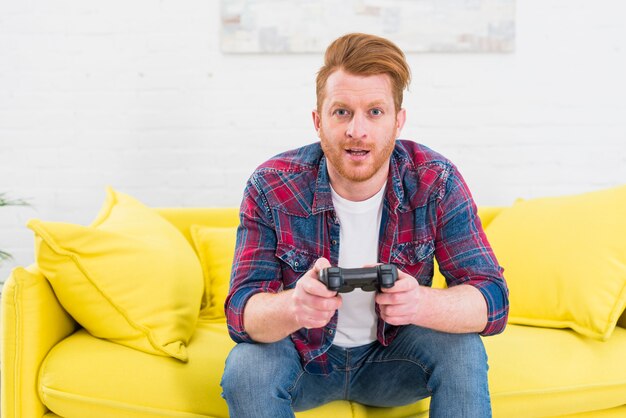 ビデオゲームで遊んで黄色のソファーに座っていた興奮している若い男の肖像