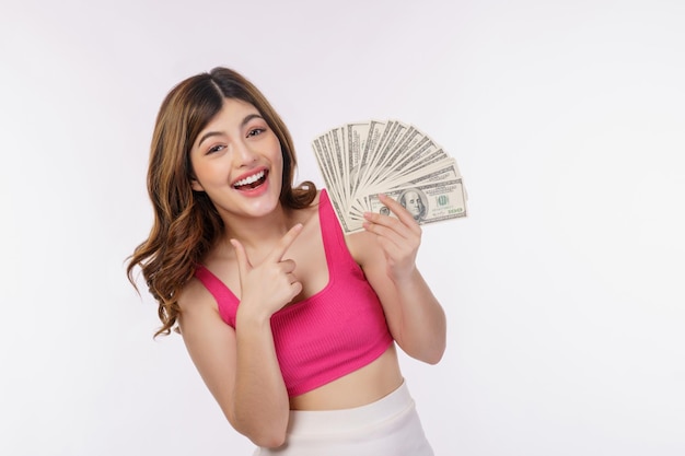 Портрет взволнованной молодой женщины, держащей кучу банкнот в долларах и указывающей пальцем на деньги, изолированные на белом фоне