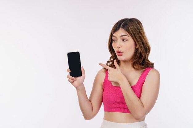 휴대폰을 들고 흰색 배경에 격리된 스마트폰을 손가락으로 가리키는 흥분한 젊은 여성의 초상화