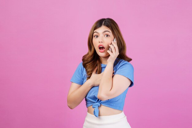 분홍색 배경 위에 격리된 휴대전화로 통화하는 캐주얼 티셔츠를 입은 흥분한 젊은 여성의 초상화