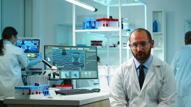 Portrait of exhausted chemist looking at camera in modern equipped lab late at night. Team of biochemists examining virus evolution using high tech and chemistry tool for research, vaccine development