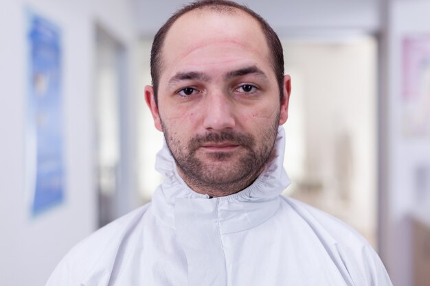 Portrait of exhausted doctor in office looking on camera wearing ppe suit without face shield sitting on chair in waiting room clinic