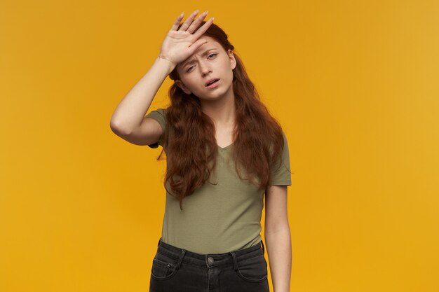 Portrait of exhausted, tired lady with long ginger hair. wearing green t-shirt. people and emotion concept. touching her forehead, feels sick. isolated over orange wall