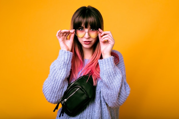 portrait of exited happy woman with unusual trendy ombre pink hairs posing at yellow wall, surprised emotions, cozy sweater and vintage sunglasses.