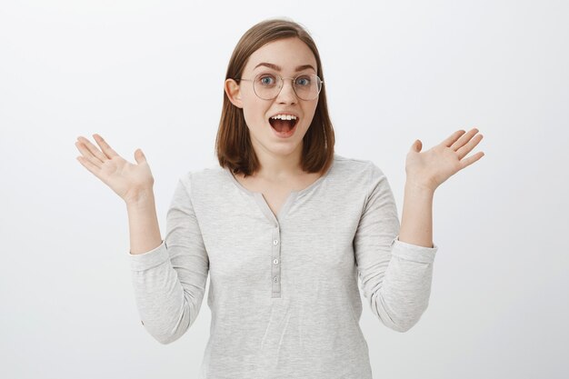 Portrait of expressive joyful smart european female in trendy glasses and white blouse talking passionately about interesting hobby gesturing with raised palms being emotional over grey wall