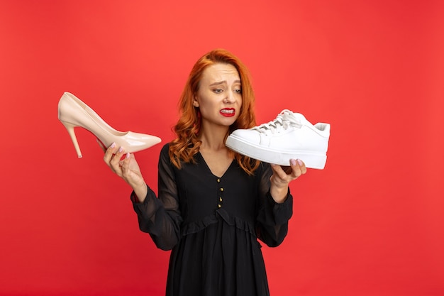 Portrait of expressive woman holding sneakers and heeled shoes isolated on red