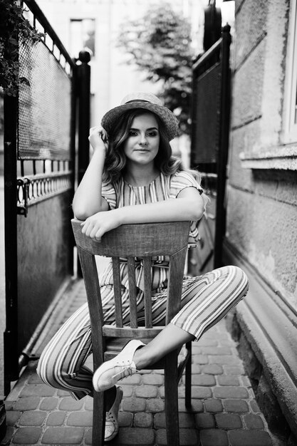 Portrait of a fabulous young woman in striped overall and hat sitting and posing on the chair outdoor Black and white photo